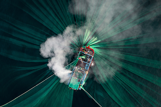 Thien Nguyen Ngoc - A fishing boat off Hon Yen as its long trail of smoke perfectly aligns with the shape of the green nets under the surface. Vietnam. 3rd place Ocean Photographer of the Year 2024